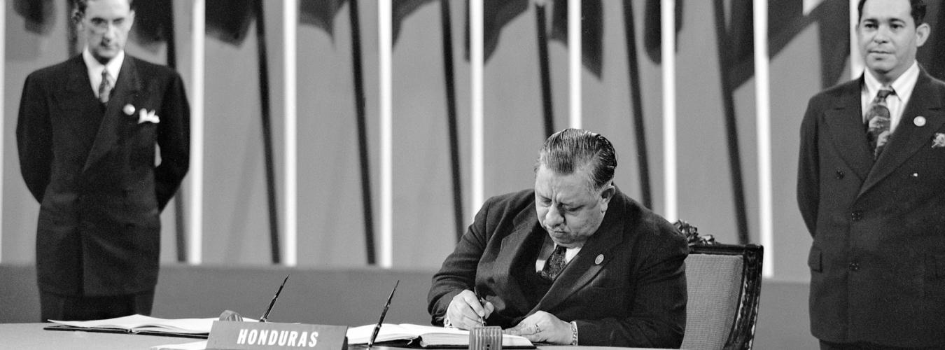 Председатель делегации Гондураса, сидящий на переднем плане, подписывает Устав ООН в 1945 году, а два члена делегации на заднем плане наблюдают.
